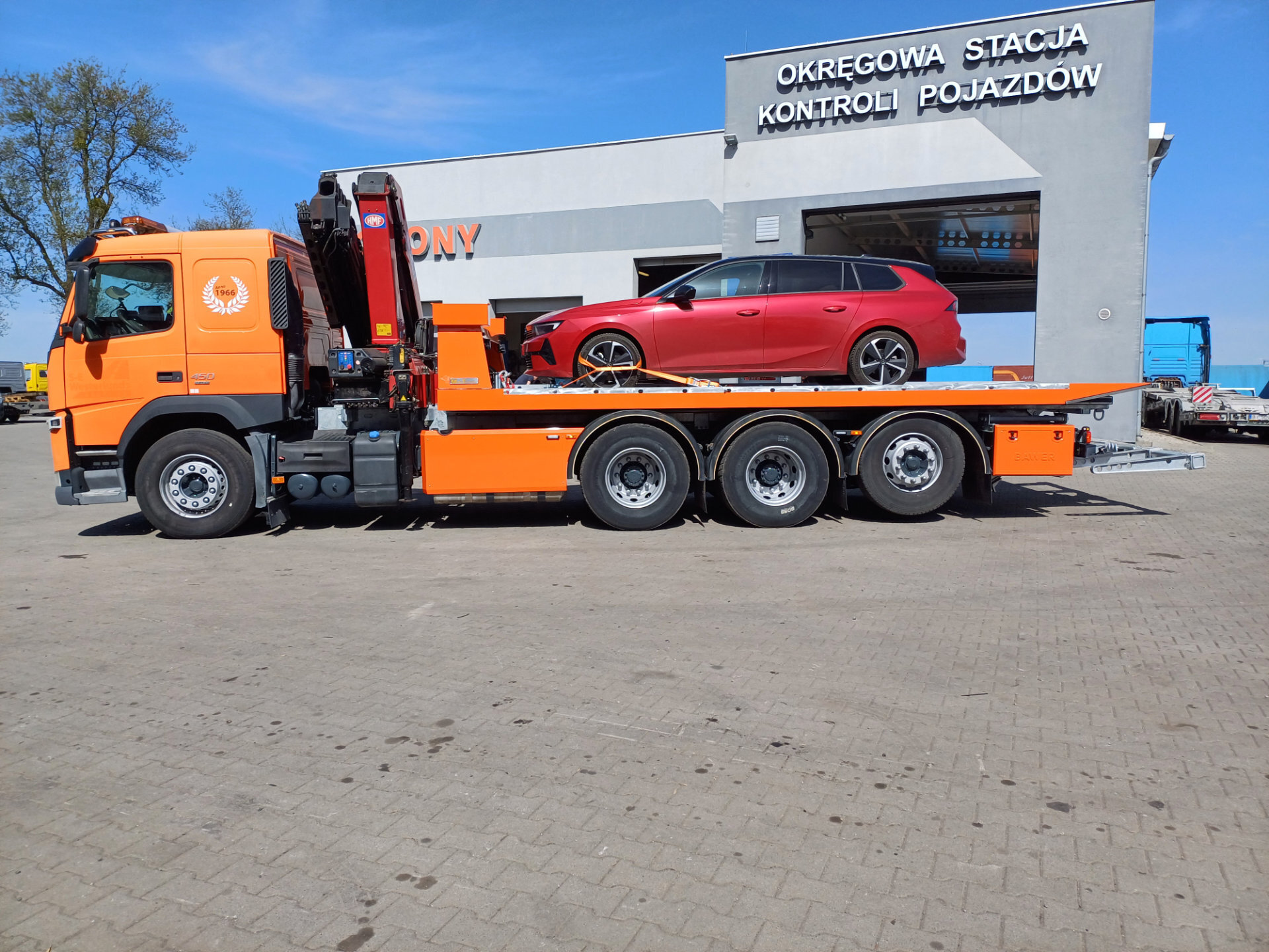 Pomarańczowy Volvo FM 450 z czerwoną osobową na platformie hydraulicznej