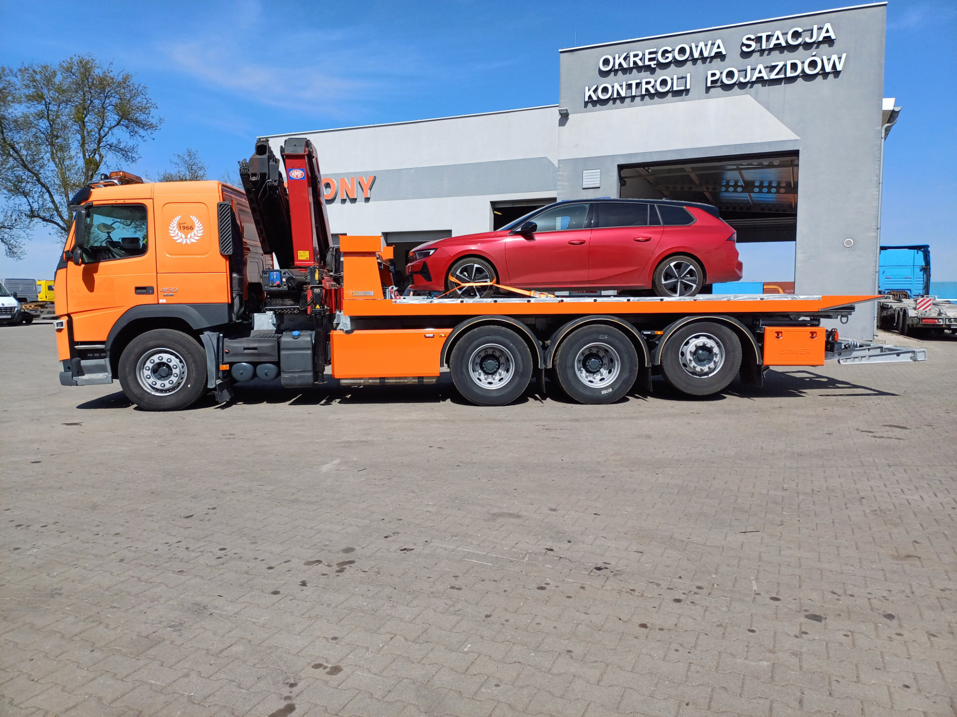 Pomarańczowy Volvo FM 450 laweta przewożąca czerwony samochód osobowy