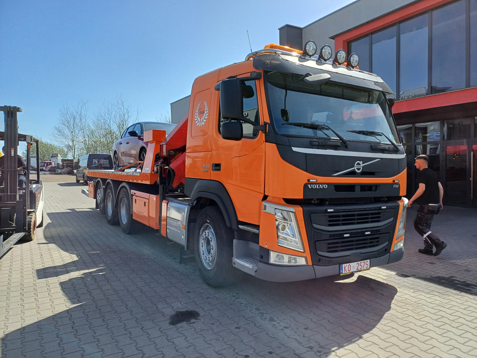 Pomarańczowy Volvo FM 450 z platformą hydrauliczną i samochodem.